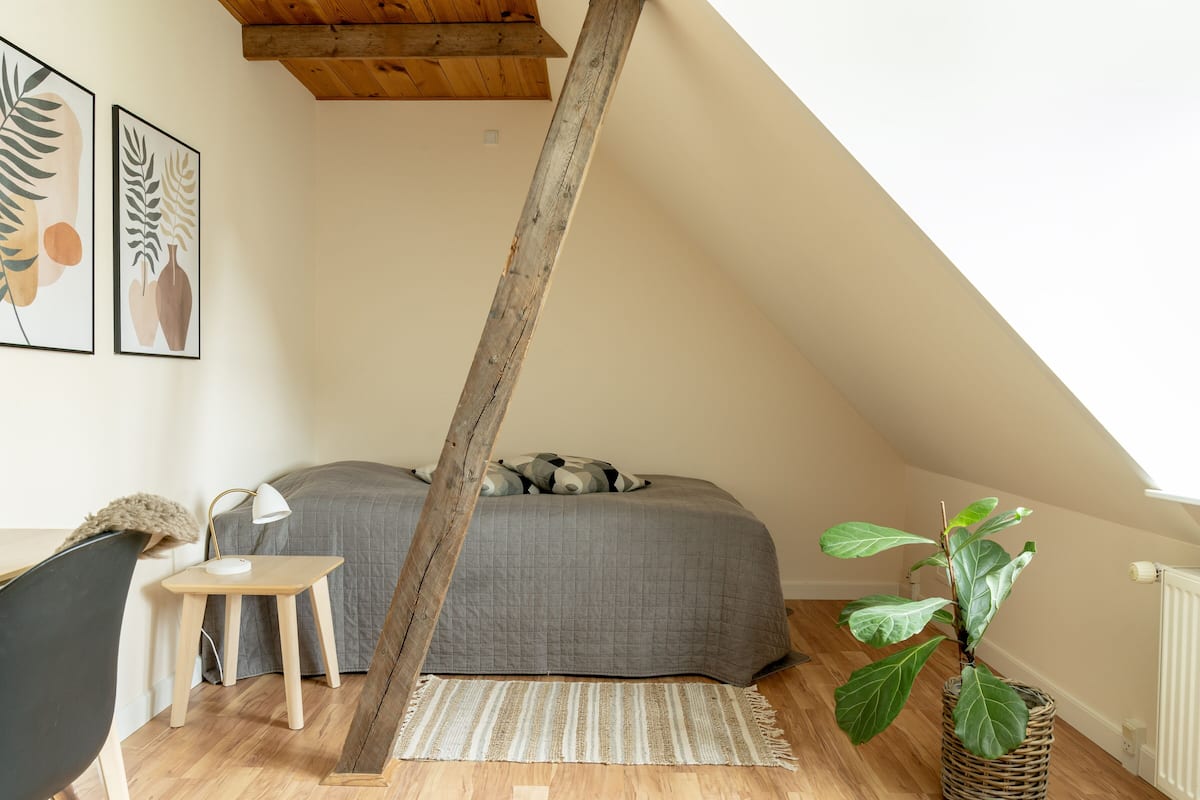 A cozy attic bedroom with a sloped ceiling, a bed with a gray quilt, a small wooden table, a chair, a plant, and two framed botanical prints on the wall.