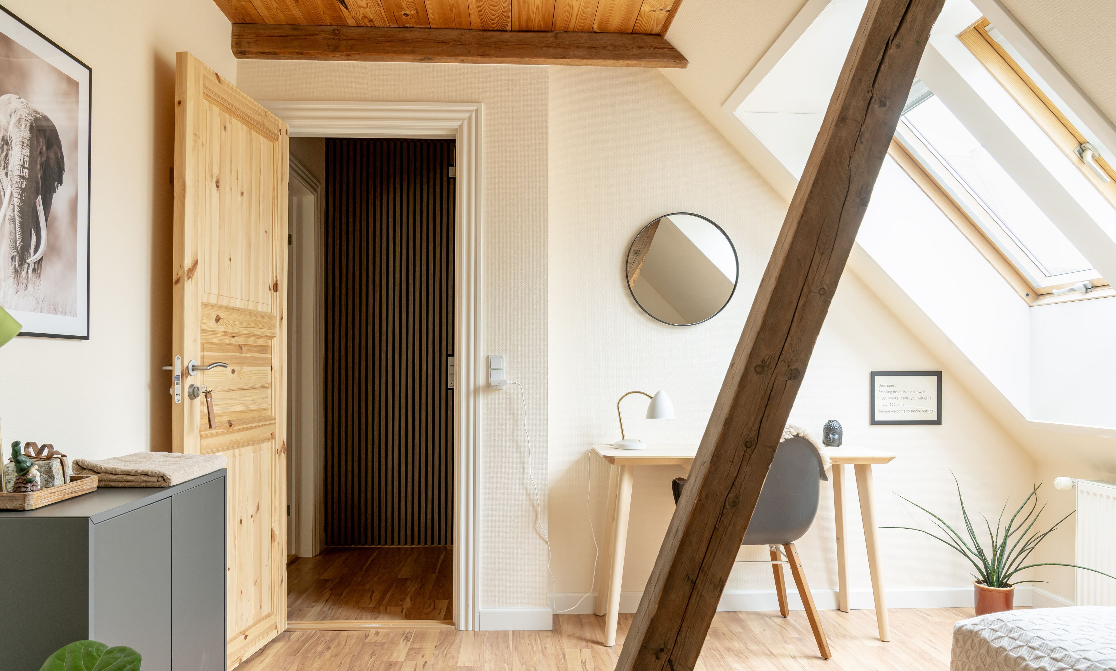 A room with a wooden ceiling, a desk with a chair, a round mirror, and a potted plant.