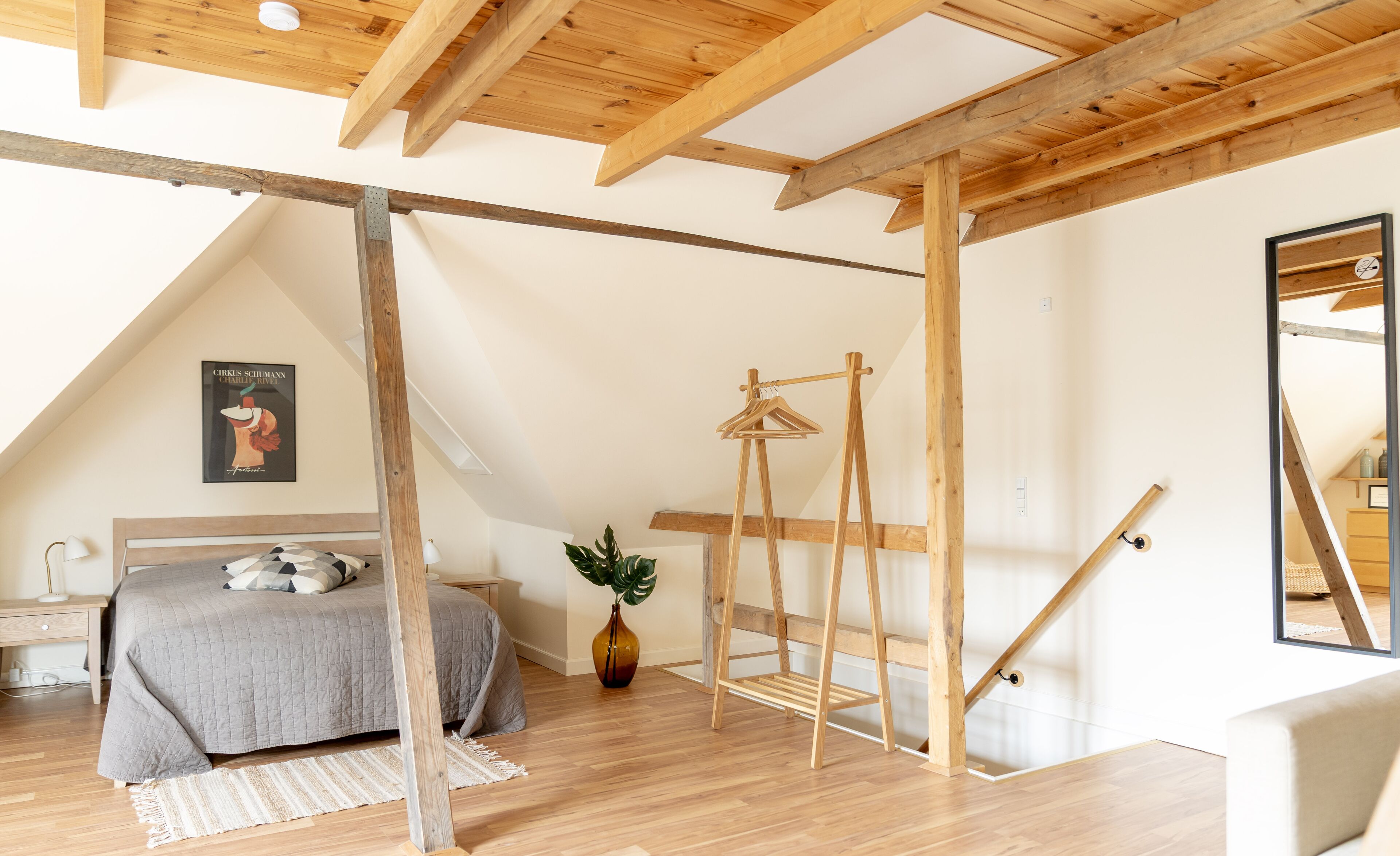 A bedroom with a sloped ceiling, wooden beams, a bed with a grey quilt, a wooden ladder, and a framed picture on the wall.