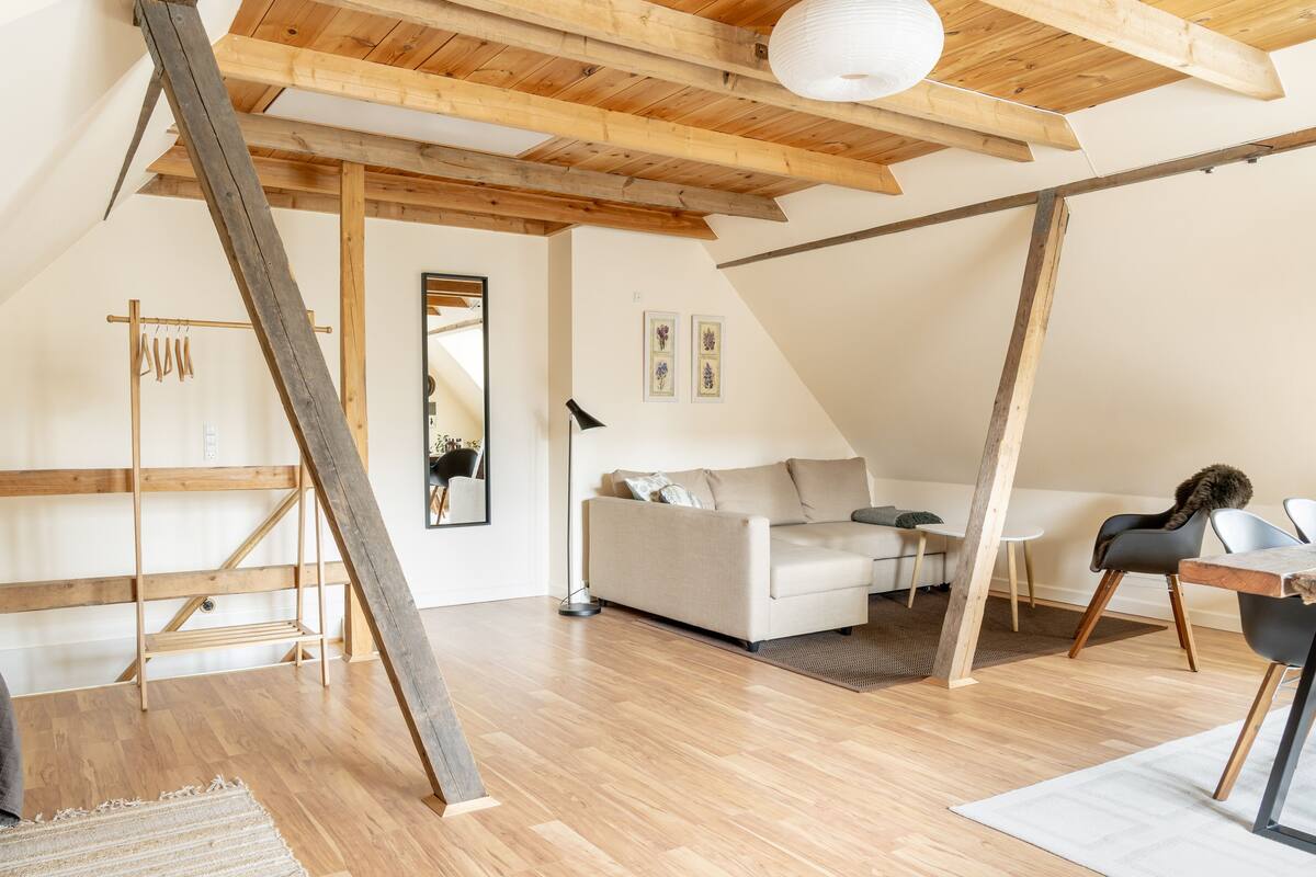 A loft-style room with exposed wooden beams, a white sofa, a dining table, and a mirror.