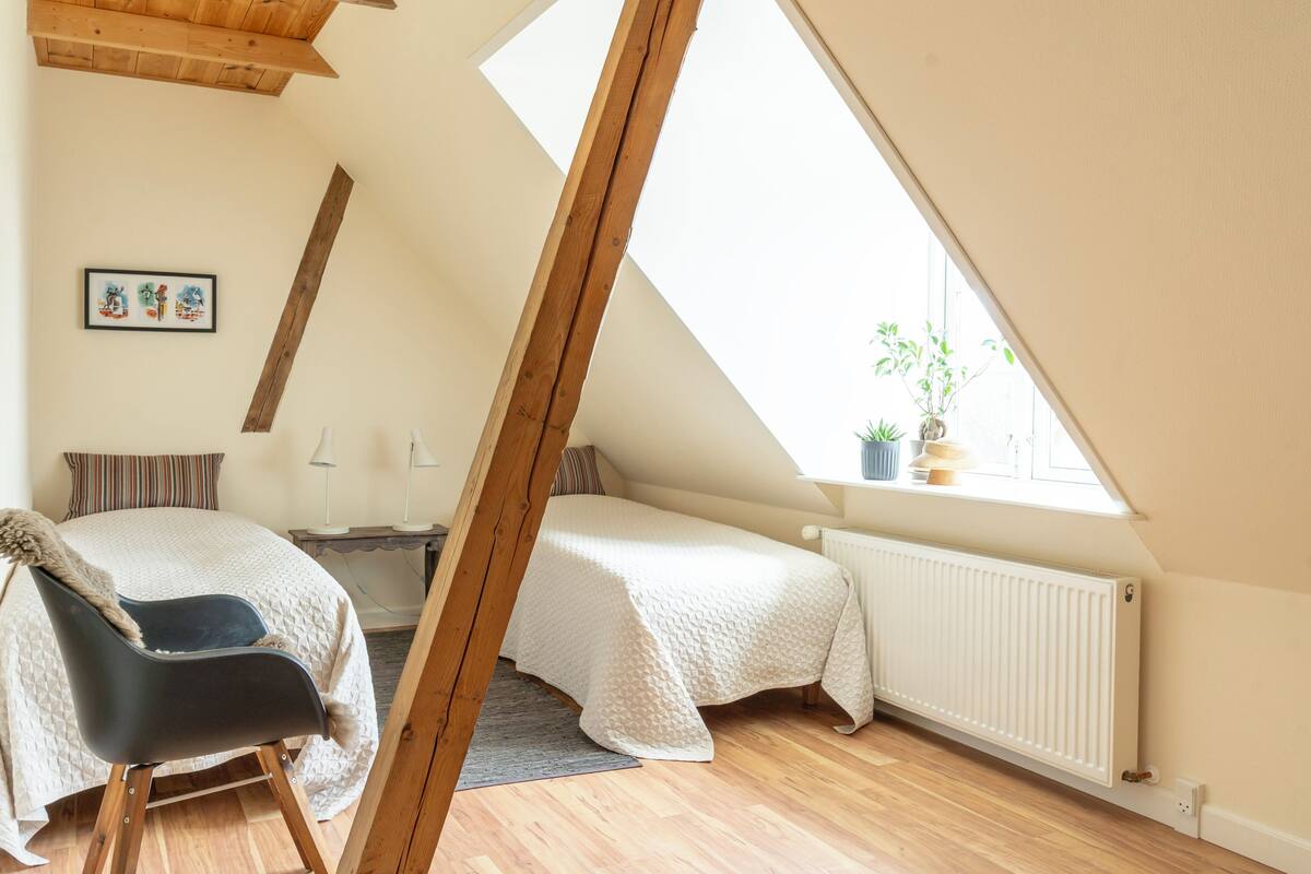 A bedroom with a sloped ceiling, a bed, a chair, a radiator, and a window with a plant.
