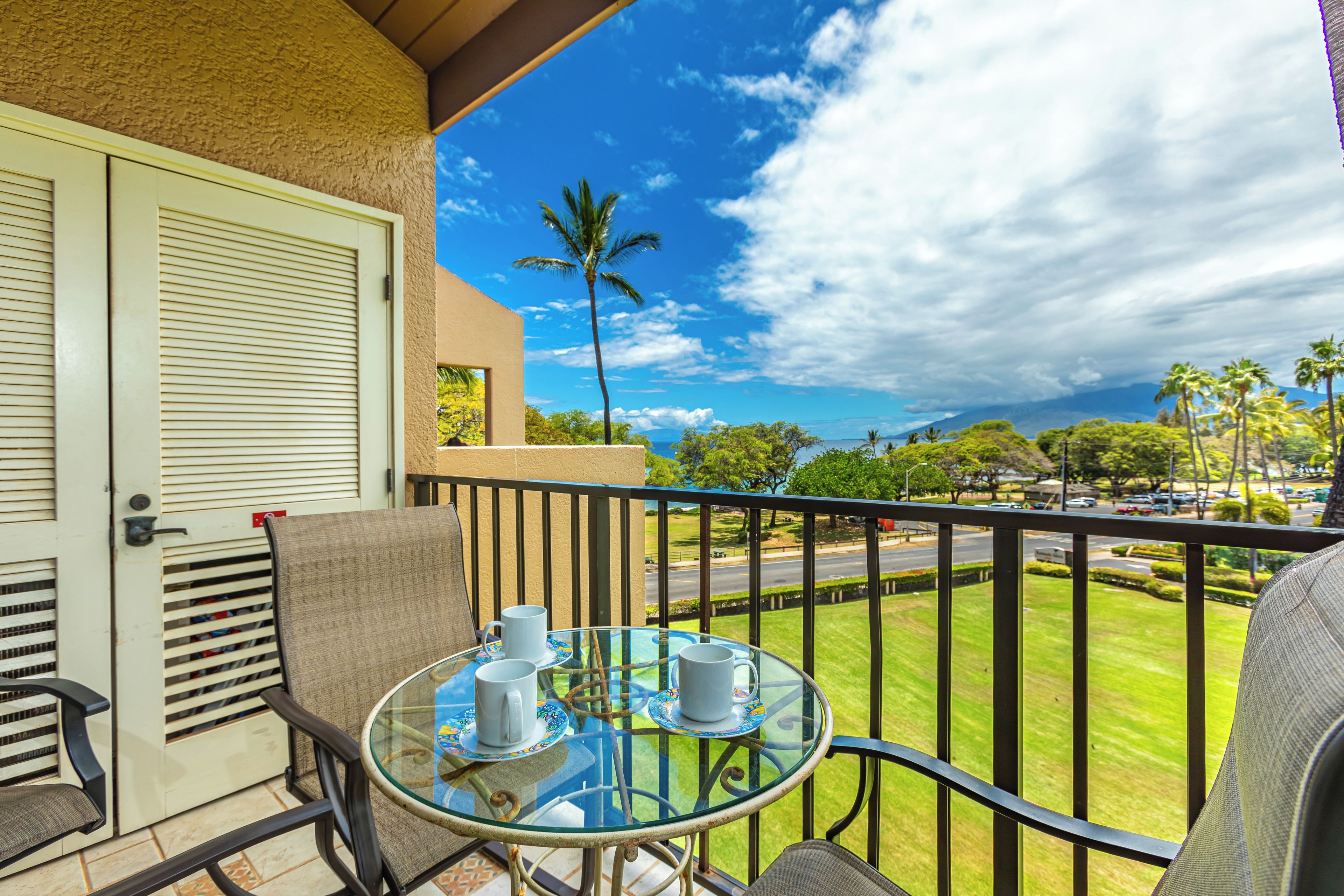A balcony with a glass table, two chairs, and a view of a green lawn and palm trees.