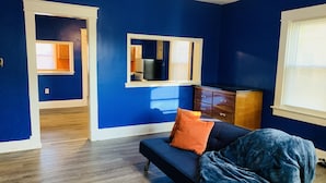A room with blue walls, a dark blue sofa with orange and blue cushions, a wooden dresser, and a mirror on the wall. - Royalty Homes - College Style 3 bedroom Apartment (Baltimore)