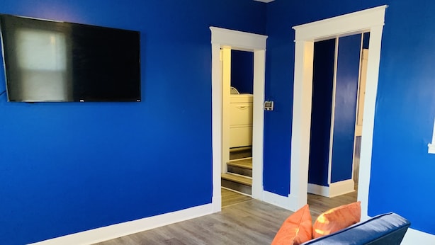 A room with blue walls, a flat-screen TV mounted on the wall, and a sofa with a blue cushion and an orange pillow. - Royalty Homes - College Style 3 bedroom Apartment (Baltimore)
