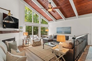 A spacious living room with a high ceiling, wooden beams, a fireplace, a television, and a large window offering a view of trees.