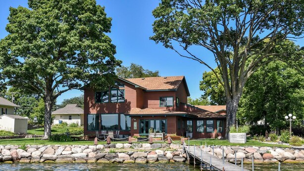 A two-story house with a dock extending into a body of water, surrounded by trees and a stone wall. - Sandy Shore Beach House:  Lake Minnetonka (Mound)