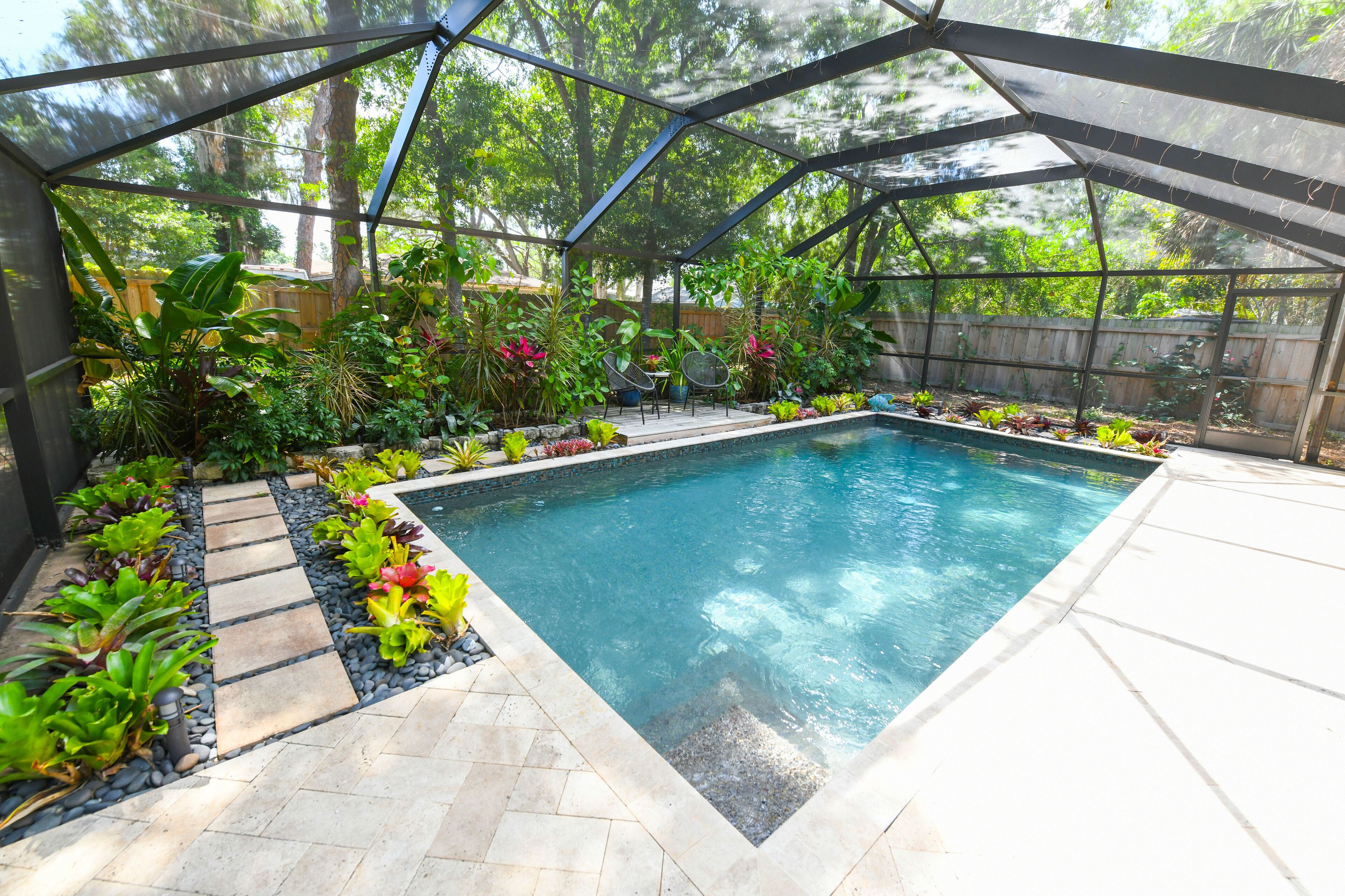 A swimming pool with a glass enclosure, surrounded by various plants and a wooden fence.