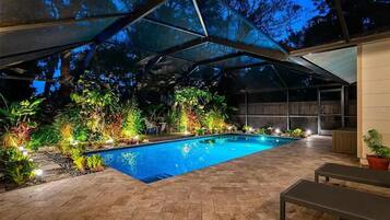 A covered outdoor swimming pool with a patio area, surrounded by lush greenery and a pergola structure.
