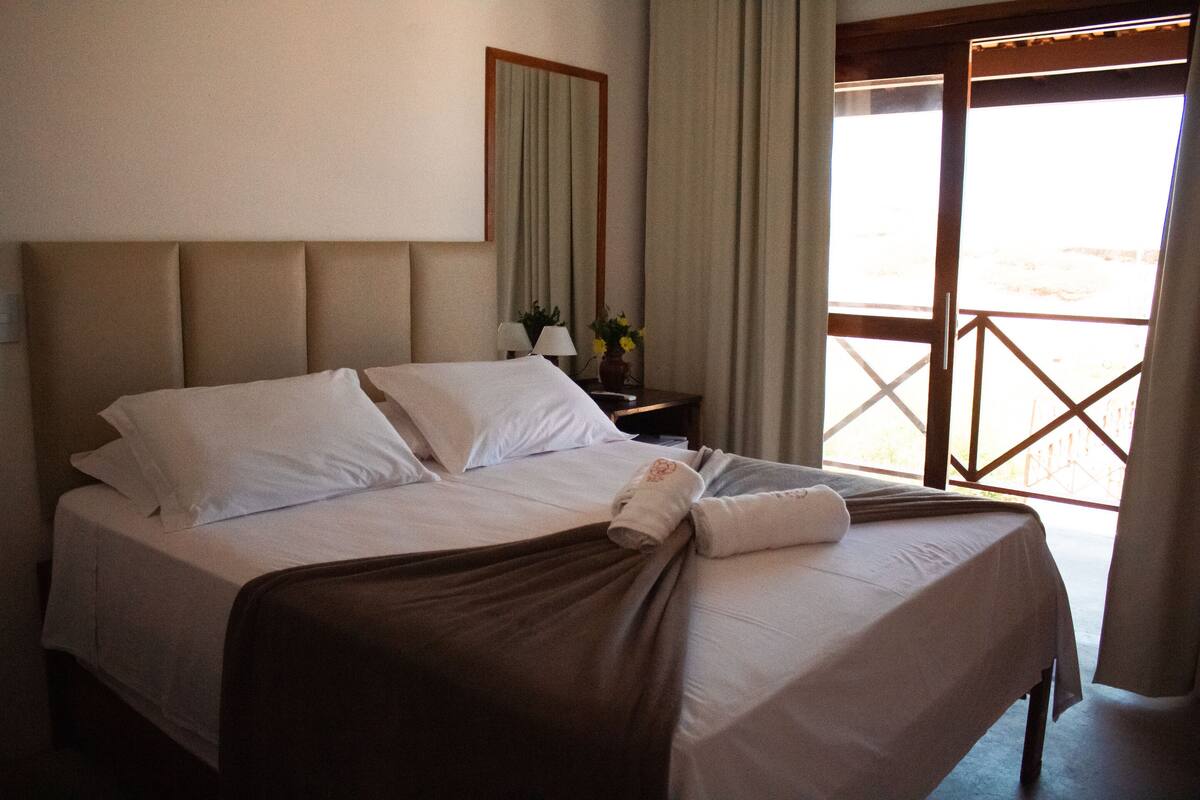 Una cama bien tendida con sábanas blancas y un plaid marrón, una mesita de noche con una lámpara y un jarrón con flores, y un ventanal con vistas al exterior.