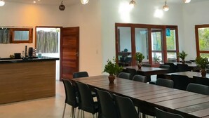 Een ruime eethoek met meerdere tafels en stoelen, een keuken met een houten aanrechtblad, en hangende bolvormige lampen.