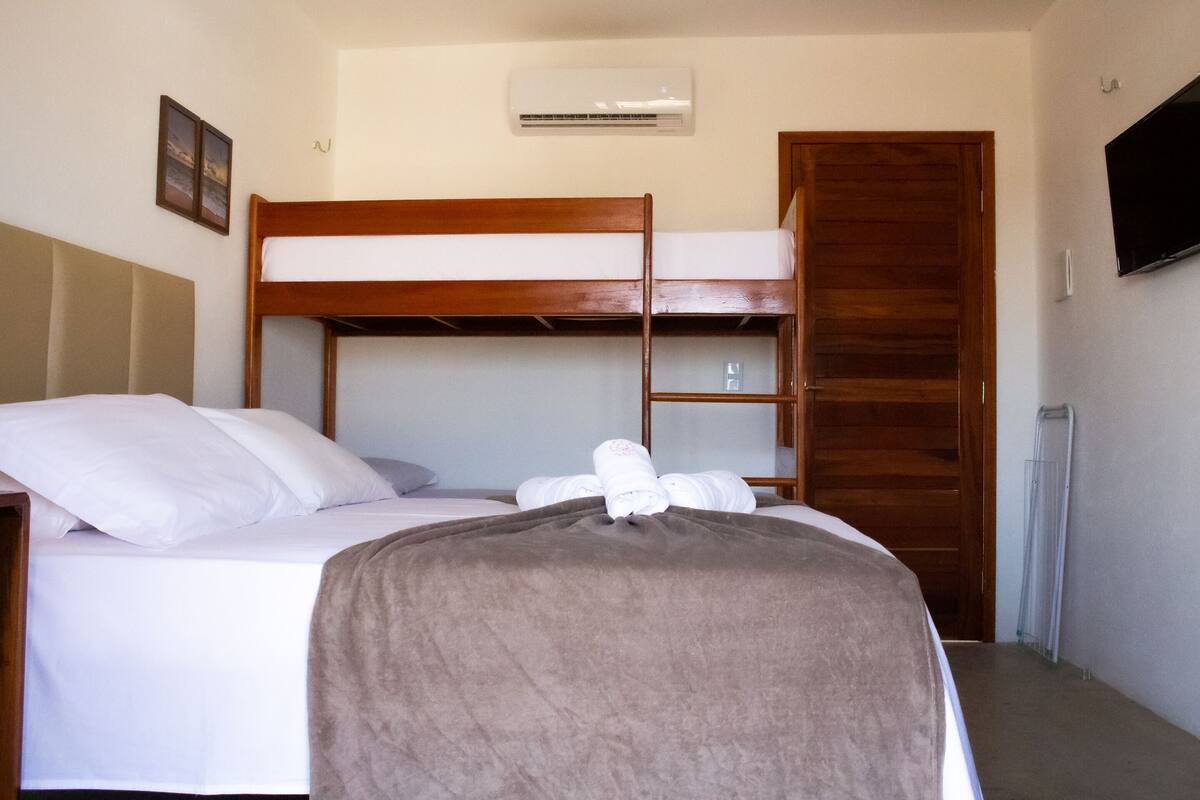 Habitación con literas, cabecera de madera, cama con almohadas, manta y televisión en la pared.