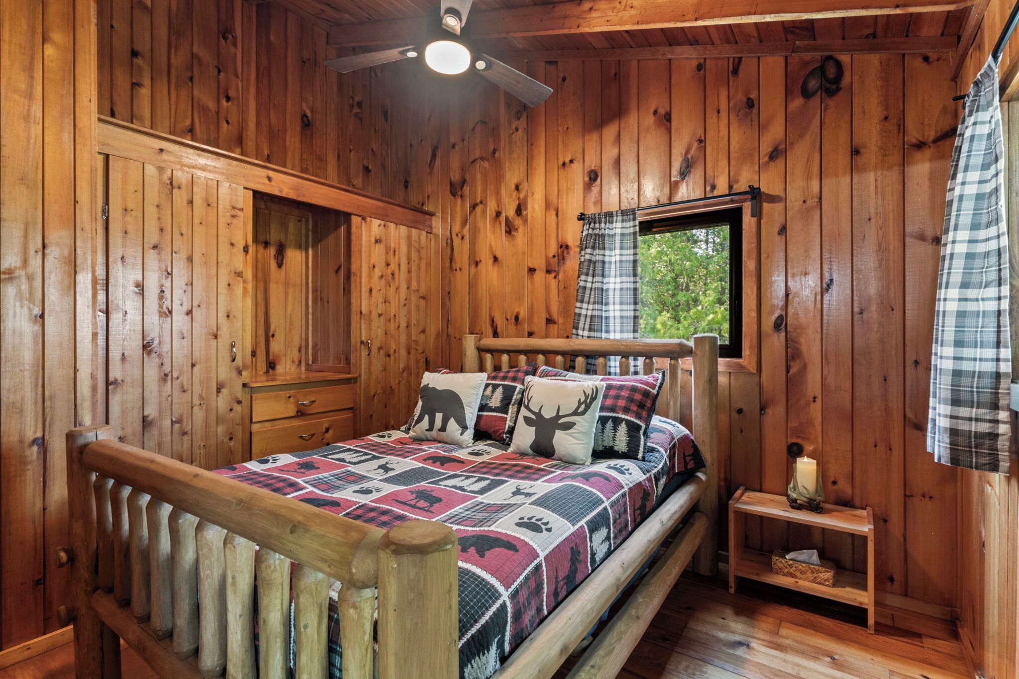 A wooden cabin bedroom with a bed, a nightstand, a window, and a ceiling fan.