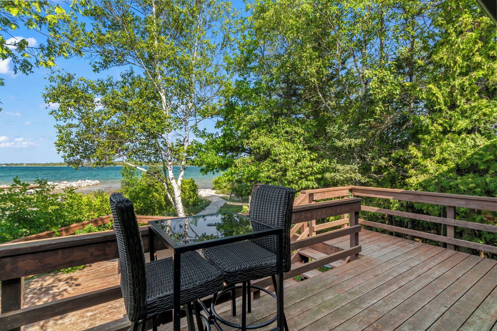A wooden deck with a glass table and black wicker chairs overlooking a beach and ocean.