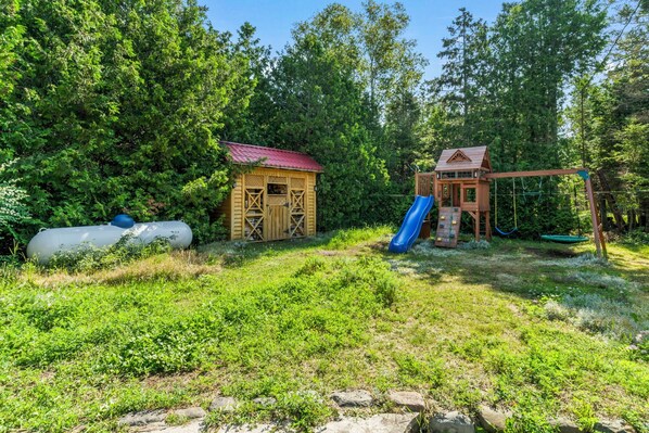 A rustic wooden playhouse with a red roof, a slide, and a swing set in a wooded area.