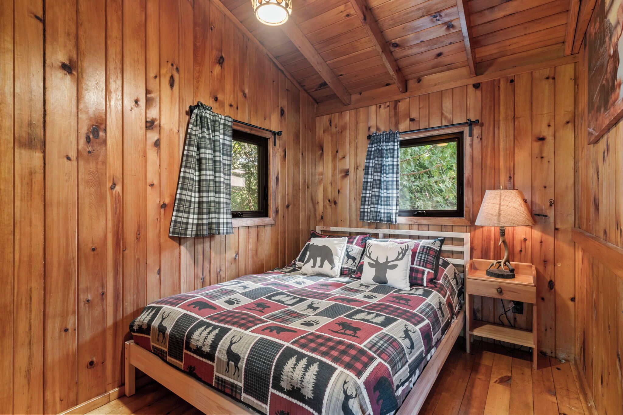 A wooden cabin bedroom with a bed, two windows with plaid curtains, a nightstand with a lamp, and a quilt with animal prints.