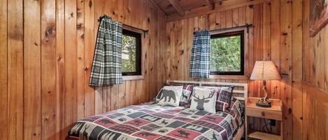 A wooden cabin bedroom with a bed, two windows with plaid curtains, a nightstand with a lamp, and a quilt with animal prints.