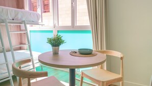 Une pièce avec un lit superposé, une table ronde en bois entourée de chaises, une plante en pot et une vue sur un immeuble par la fenêtre.