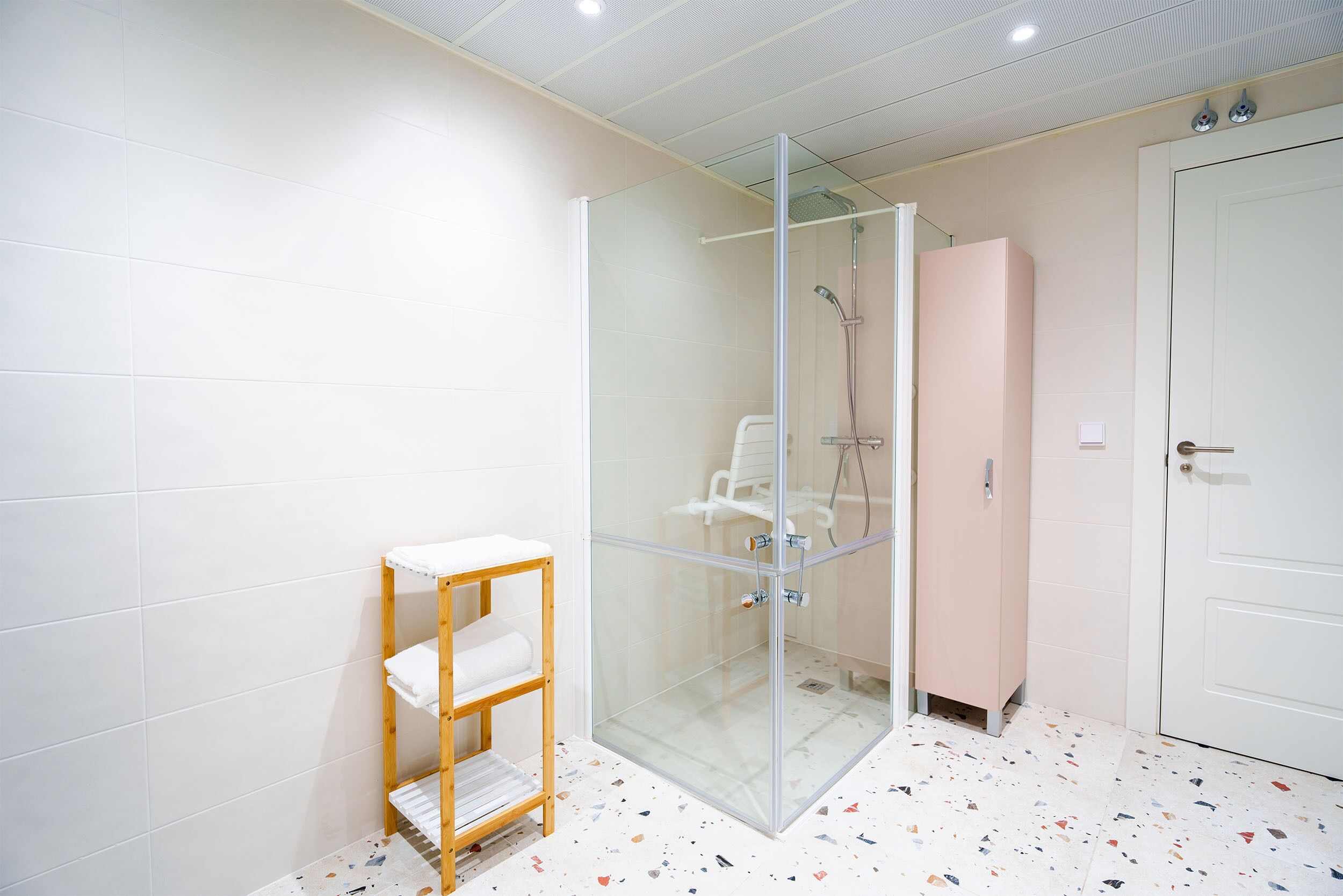 A modern bathroom with a glass-enclosed shower, a wooden shelf with towels, and a white door.