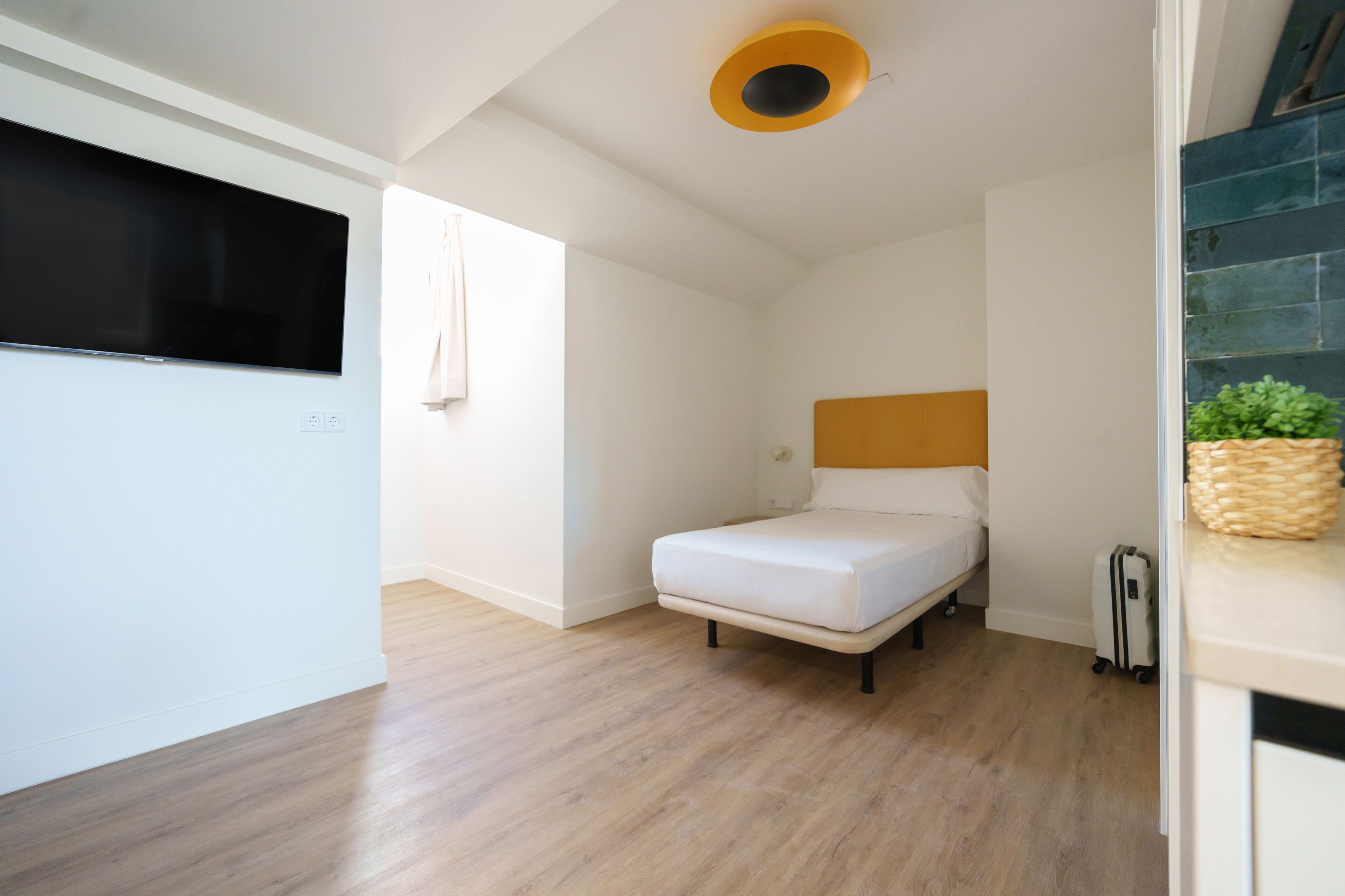 A compact room with a bed, a small bedside table, a suitcase, and a ceiling light fixture.