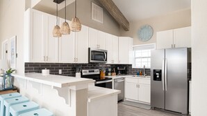 A modern kitchen with white cabinets, a stainless steel refrigerator, a microwave, and a sink. There are three blue bar stools and a hanging light fixture. - Beach Bliss | Epic Views, Games & Pets! (Crystal Beach)