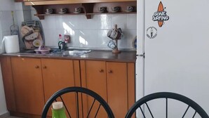 A kitchen with wooden cabinets, a white refrigerator with a sticker, and a table with a green vase and blue plates. - Marilia House Skiathos (Skiathos)