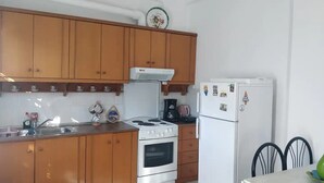 A compact kitchen with wooden cabinets, a stove, a refrigerator, and a dining area with chairs. - Marilia House Skiathos (Skiathos)
