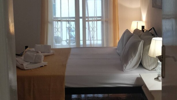 A bedroom with a bed, a bedside table with a lamp, a window with curtains, and a view of the outside. - Marilia House Skiathos (Skiathos)