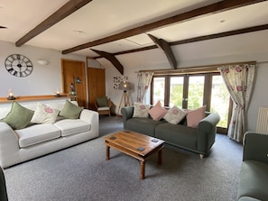 A cozy living room with a white sofa, a green sofa, and a wooden coffee table. There is a large clock on the wall and a window with curtains. - Foxglove Barn (South Brent)