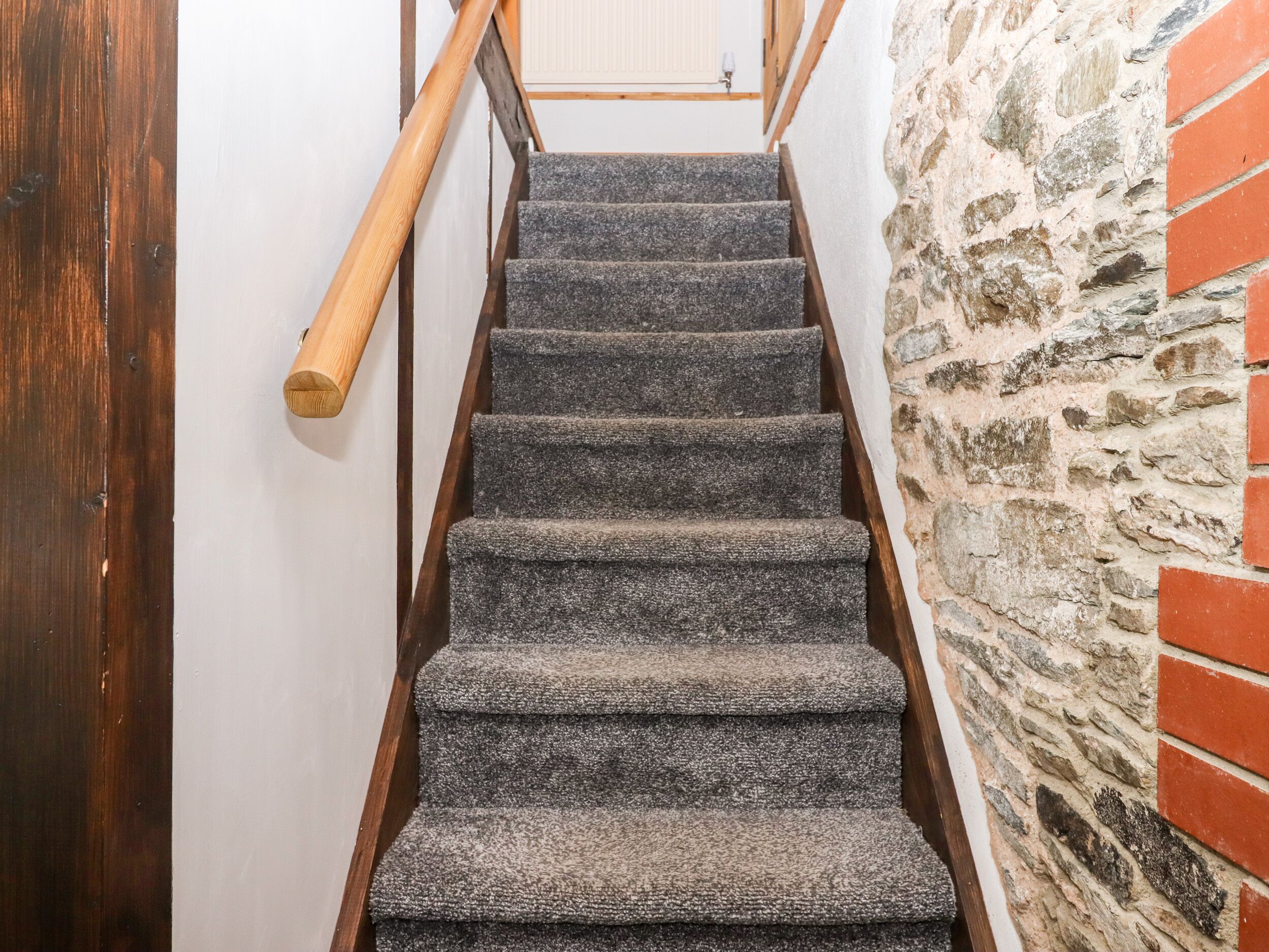 Un escalier avec des marches recouvertes de moquette et des rampes en bois, menant à une porte au-dessus de laquelle se trouve un interrupteur.