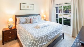 A neatly made bed with a patterned bedspread, two bedside tables with lamps, a framed artwork above the bed, and a window with curtains. - Screened Pool & Themed Rooms 8 Miles to Disney (Kissimmee)