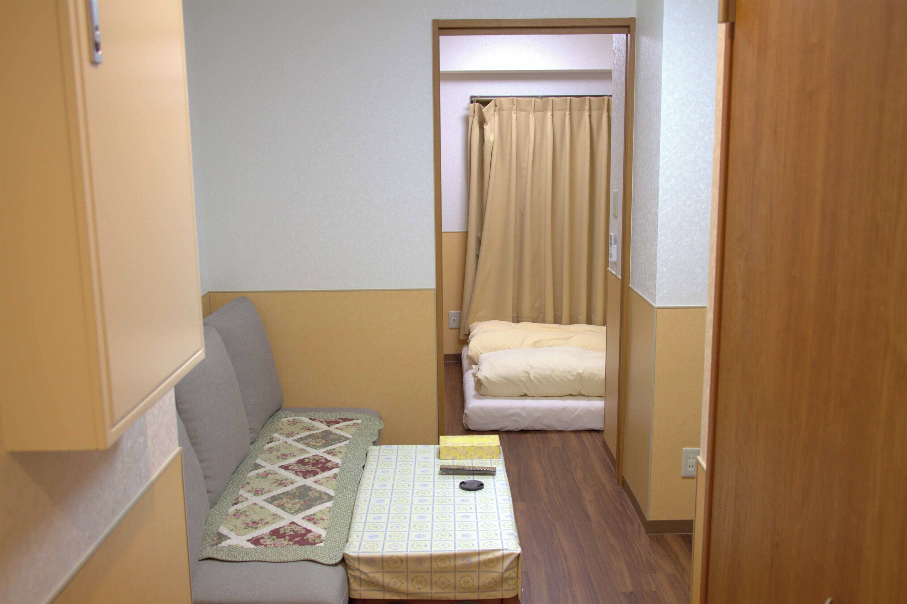 A compact room with a sofa, a small table with a quilt, a bed with a beige blanket, and a curtain.