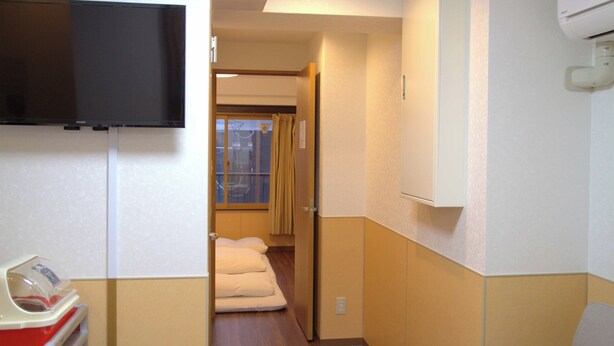 A hotel corridor with a television mounted on the wall, a bed with white bedding, and a window with curtains. - Ueno House VIP (Tokyo)