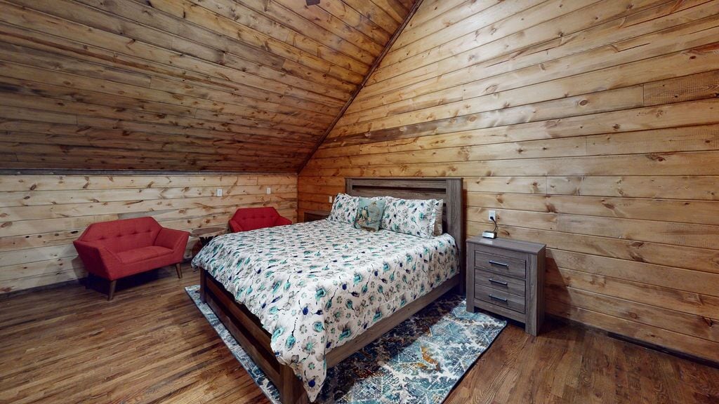 A bedroom with a wooden sloped ceiling, a bed with floral bedding, a red chair, a nightstand, and a small dresser.