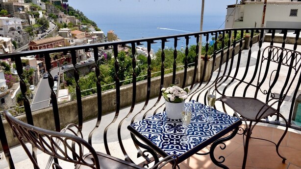 Un balcon avec une table et des chaises, donnant sur une ville côtière avec ses bâtiments et la mer.