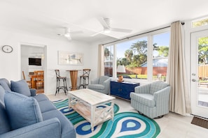 A spacious living room with a large sofa, a coffee table, and two armchairs. There is a dining area with a table and chairs, and a view of the backyard through large windows.