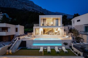 A modern two-story house with a swimming pool, outdoor seating, and a mountain backdrop.