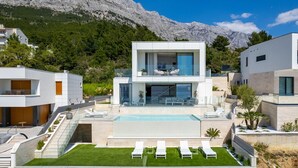 Ein modernes, zweistöckiges Haus mit Swimmingpool, Sitzgelegenheiten im Freien und einer Bergkulisse.