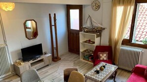 Uma sala de estar com televisão, uma escada de madeira, uma estante com objetos decorativos, um sofá vermelho e uma janela com cortinas.