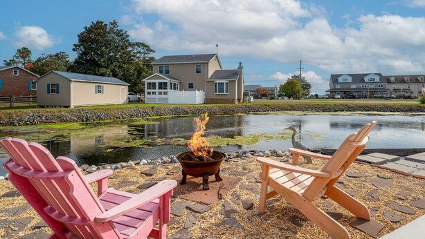 A lakeside patio with Adirondack chairs, a fire pit, and a view of residential buildings.