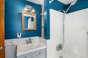 Une salle de bain avec un lavabo blanc, un miroir avec un cadre en bois et une douche équipée d’un pommeau de douche à main.