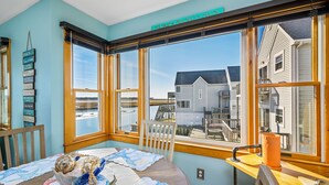 A room with large windows overlooking a beach, a wooden table with a quilt, and a sign above the windows reading "SURF SAND SUN". - Wonderview 208 by Seaside Vacations & Sales (Chincoteague Island)