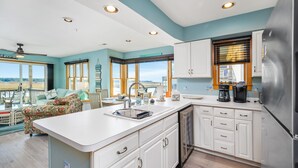 A modern kitchen with white cabinets, a stainless steel refrigerator, and a large island with a sink. There are multiple windows providing views of the outdoors. - Wonderview 208 by Seaside Vacations & Sales (Chincoteague Island)