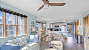 A spacious living room with a sofa, armchair, and wicker coffee table. A kitchen is visible in the background with a sign reading "CHINCHIEAU." - Wonderview 208 by Seaside Vacations & Sales (Chincoteague Island)