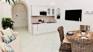 A modern kitchen with white cabinets, a built-in oven, and a large window. There is a dining area with wicker furniture and a flat-screen TV mounted on the wall. - Tyrrhenian Sea View Apartment (Campania)