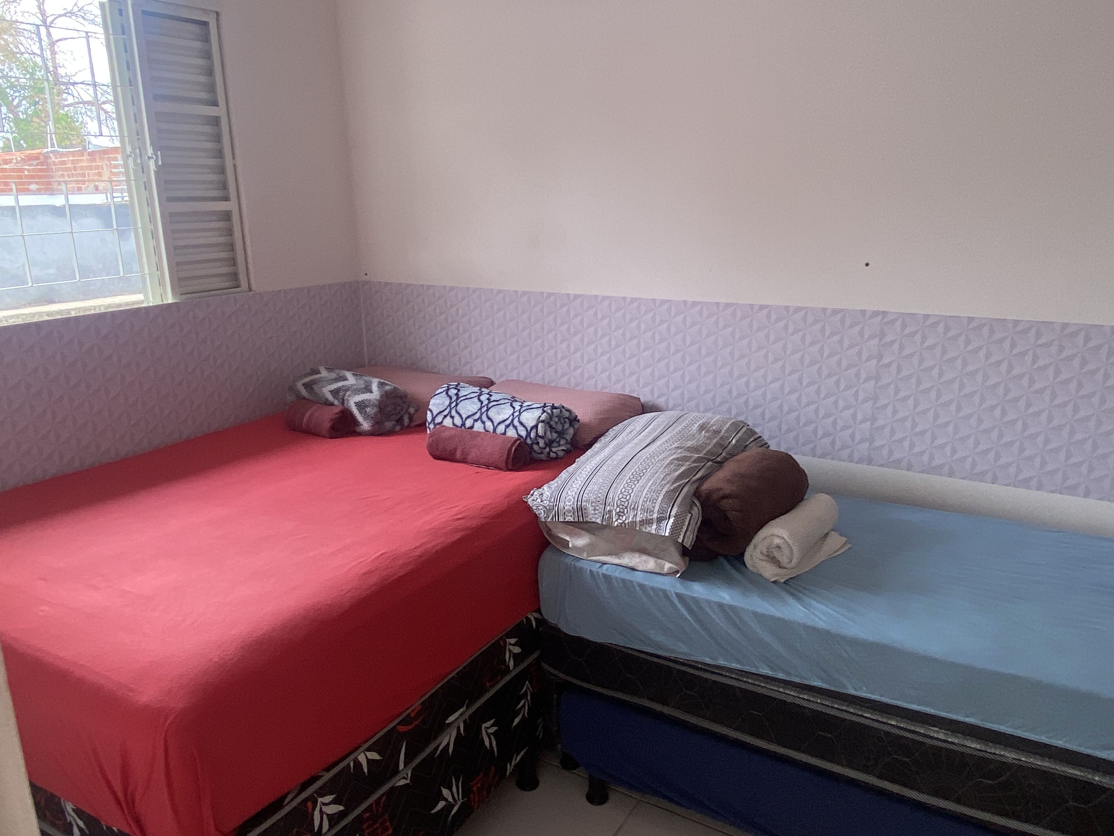 A small, single-bed room with a red and blue mattress, pillows, and a patterned blanket.