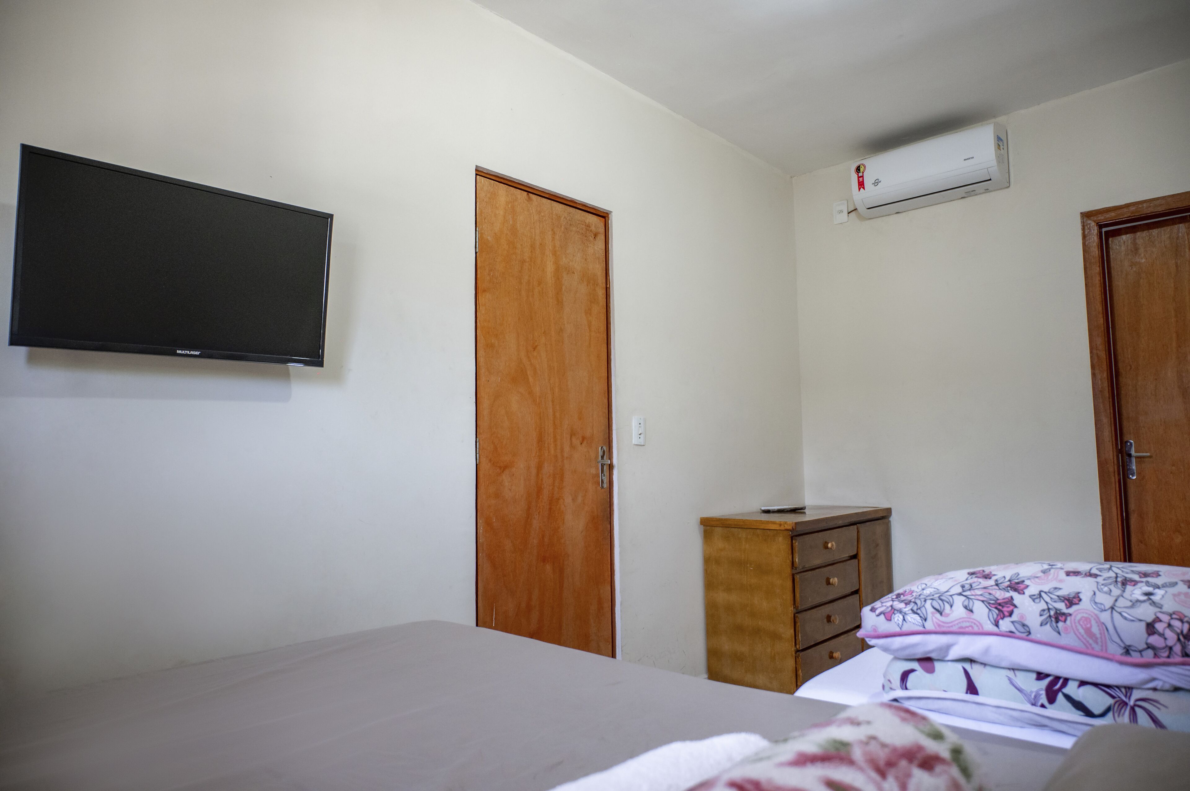 Une chambre à coucher comprenant un lit, une armoire en bois, une petite commode, un téléviseur fixé au mur et un appareil de climatisation.