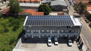 Un edificio con tetto piano ricoperto di pannelli solari, un'area di parcheggio con tre automobili bianche e un contesto urbano circostante con altri edifici e vegetazione.