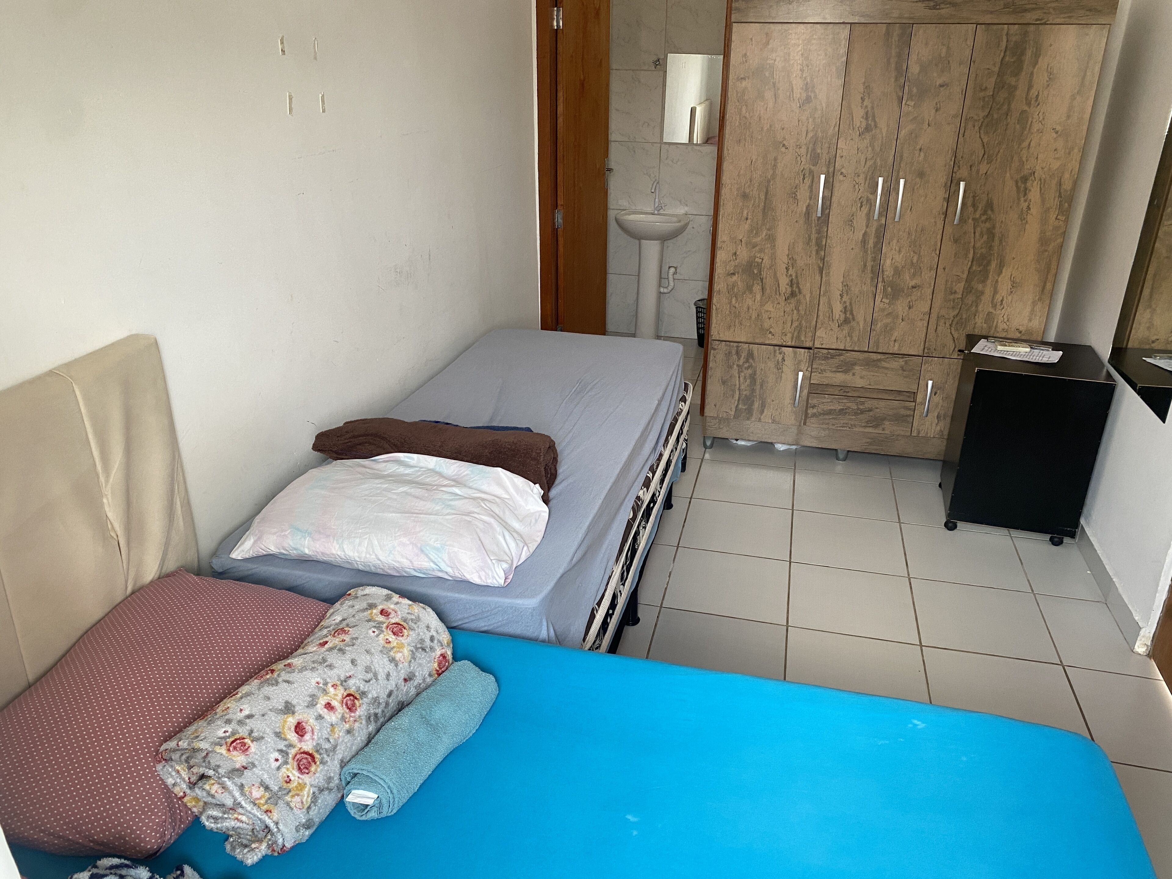 Una habitación pequeña y funcional con una cama individual, un armario de madera, un lavamanos pequeño y un mueble negro.