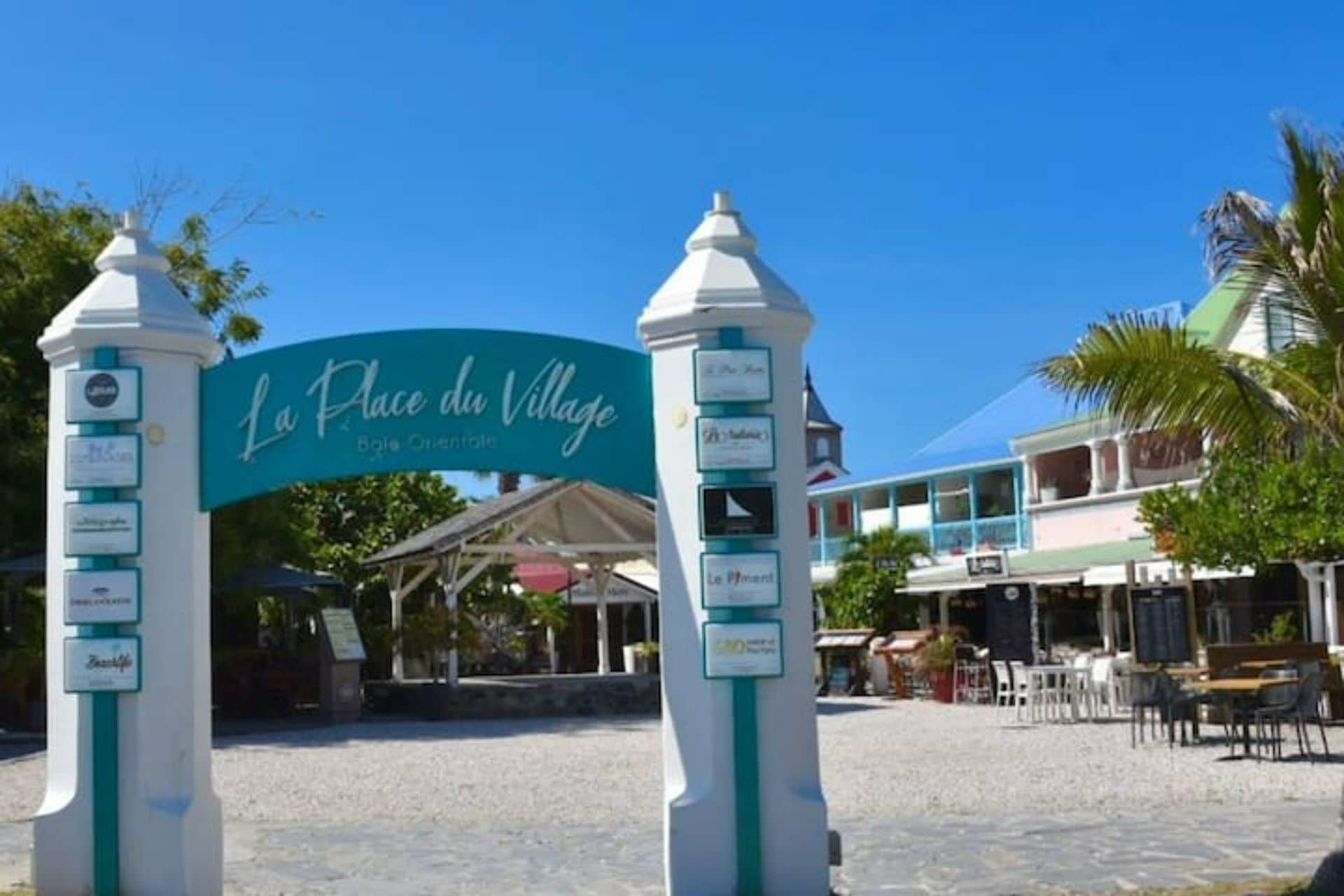 A sign for 'Place du Village' with a beach umbrella and outdoor seating area in the background.