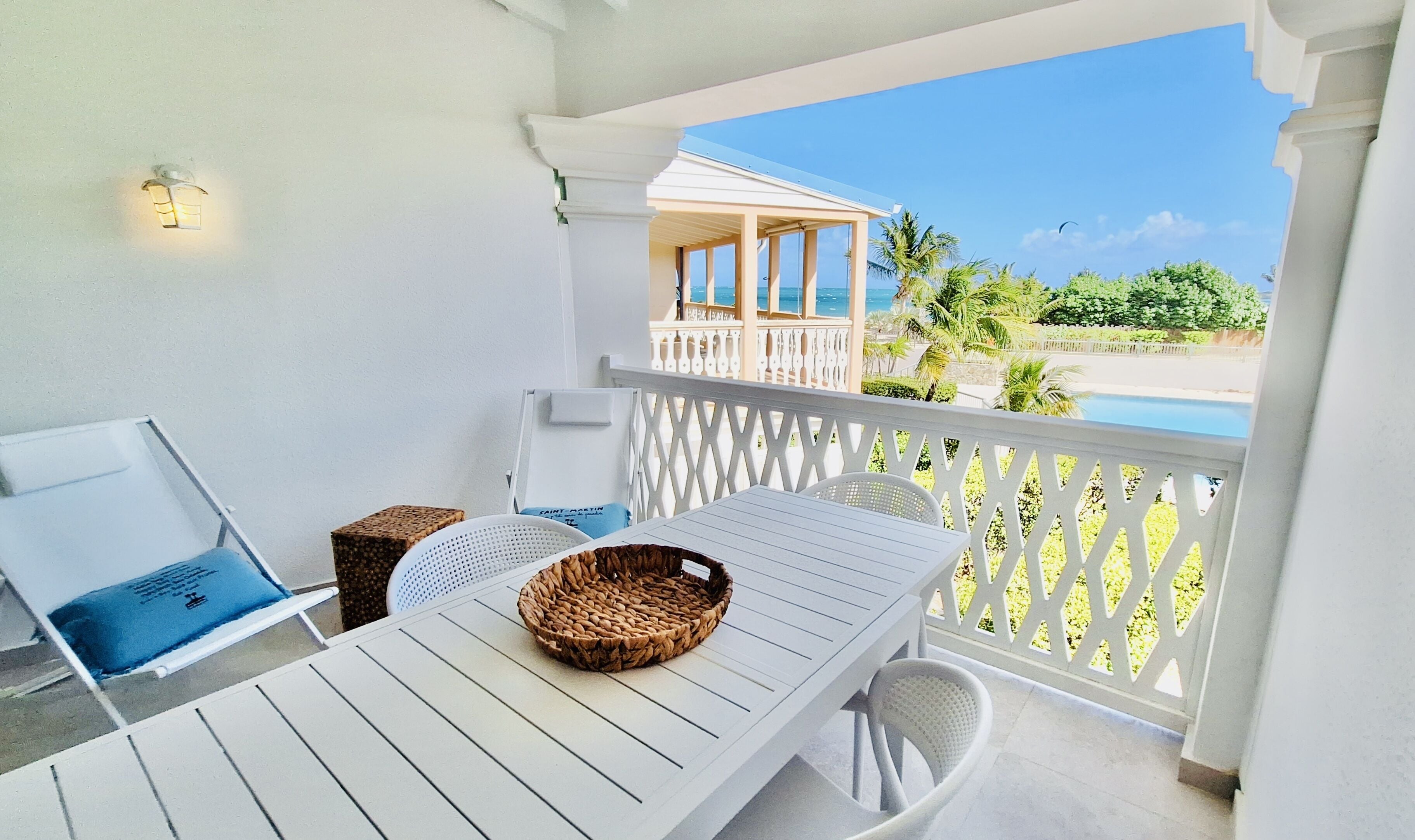 Un balcón con una mesa blanca, una cesta de mimbre y vista a una playa y palmeras.